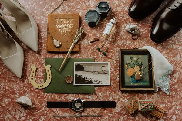 Wedding flat lay of bridal and groom shoes, ring boxes, jewelry, pocket watch, and perfume arranged on a pink marbled surface