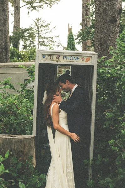 Wedding kiss as bride and groom kiss in a phone booth, her veil draped over his black suit with ivy and stone wall behind