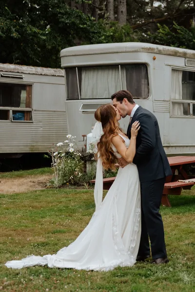 Wedding kiss as bride holds groom’s face, long veil flowing beside a vintage camper trailer on a lawn with wildflowers and trees