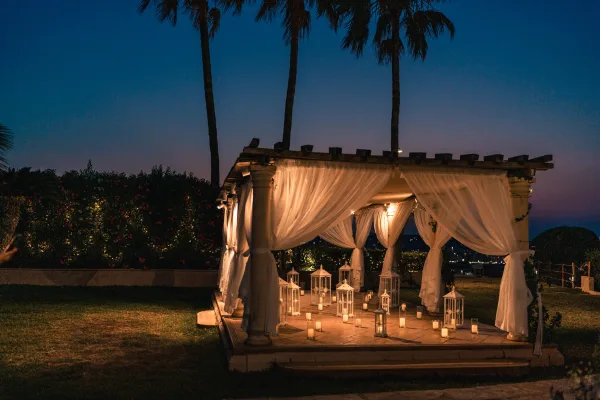 Ceremony setup for a gazebo ceremony with glowing string lights, framed by palm trees and a night sky over a manicured lawn.