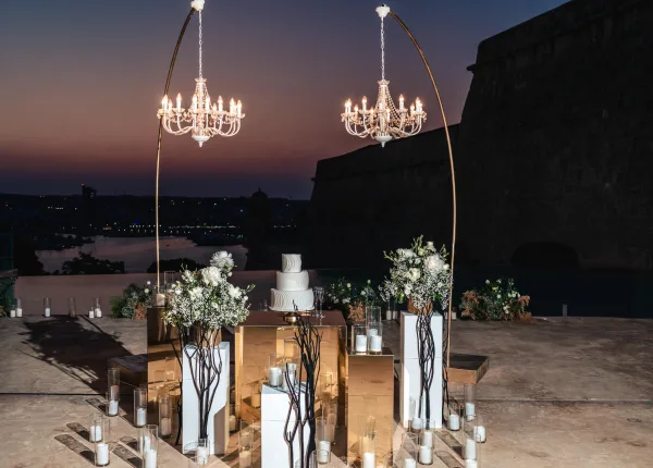 Wedding cake display with modern cake table decor, highlighted by crystal chandeliers on a waterfront terrace at sunset with city lights.