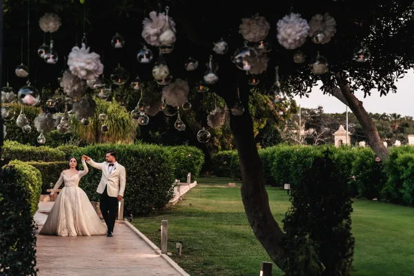 Couple portrait as the bride and groom twirl on a stone pathway under garden trees, with hanging glass orbs glowing against the evening sky.