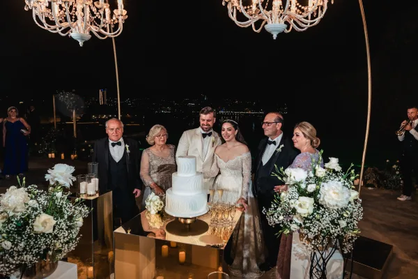 Cake cutting moment with a white tiered wedding cake on a gold cake stand, on an outdoor terrace with city lights reflecting over the waterfront.