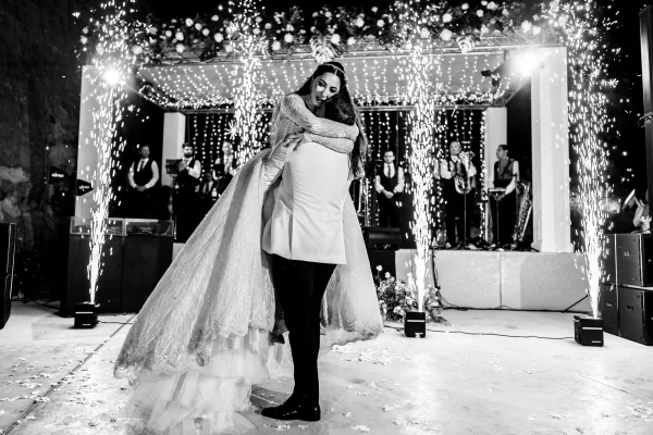 Wedding first dance with the groom lifting the bride beneath indoor sparklers, set on a stage platform framed by columns and a lighting rig.