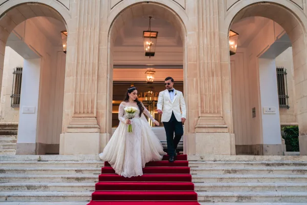 Couple portrait of a bride and groom walking down a red-carpeted staircase beneath stone arches and columns of a grand building.