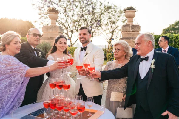 Wedding toast during cocktail hour cheers as guests raise glasses by a champagne tower on an outdoor terrace framed by garden trees at dusk.