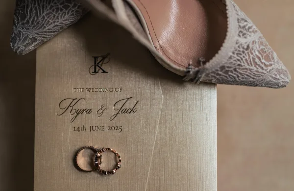 Wedding invitation flat lay featuring a monogram wedding invitation and wedding rings on soft beige fabric