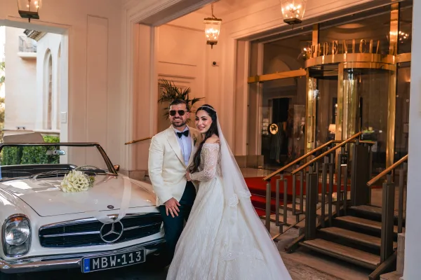 Couple portrait as a wedding car photo at a grand hotel entrance with red carpet and columns, posed beside a gleaming classic car.