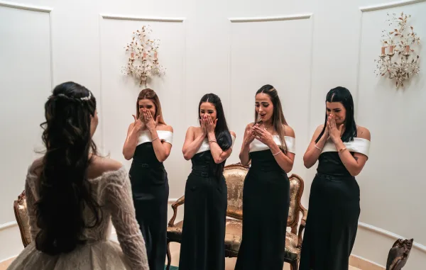 Bridesmaids first look during a bride reveal moment as they react to her lace bridal gown before a white paneled wall.