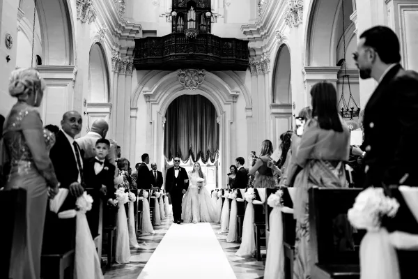 Wedding processional with the bride walking down the aisle, holding a bouquet, escorted by her father beneath vaulted arches in a grand church.