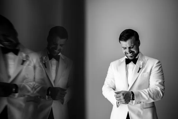 Groom getting ready adjusts his cufflinks in a candid groom portrait, standing by a mirror against a plain wall before the ceremony.