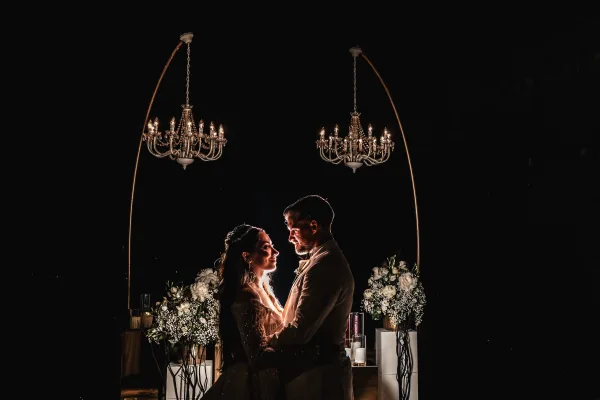 Couple portrait of a bride and groom embrace under a gold arch, dramatically lit against a black backdrop, capturing their romantic moment.