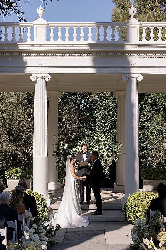 Wedding vows as bride and groom hold hands in a sunlit outdoor wedding ceremony beneath a floral arch at a white columned pavilion
