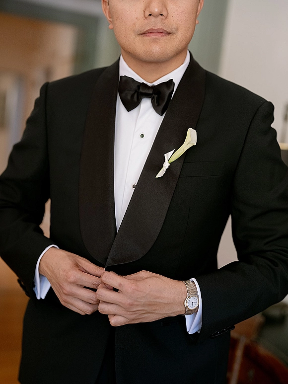 Groom portrait in a black tuxedo look with bow tie, boutonniere, and wristwatch, standing by a doorway against a neutral wall indoors