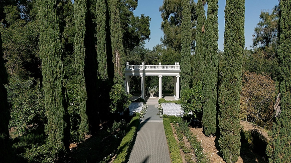 Outdoor ceremony setup with garden wedding ceremony seating, white chairs and aisle floral arrangements along a paved walkway under cypress trees
