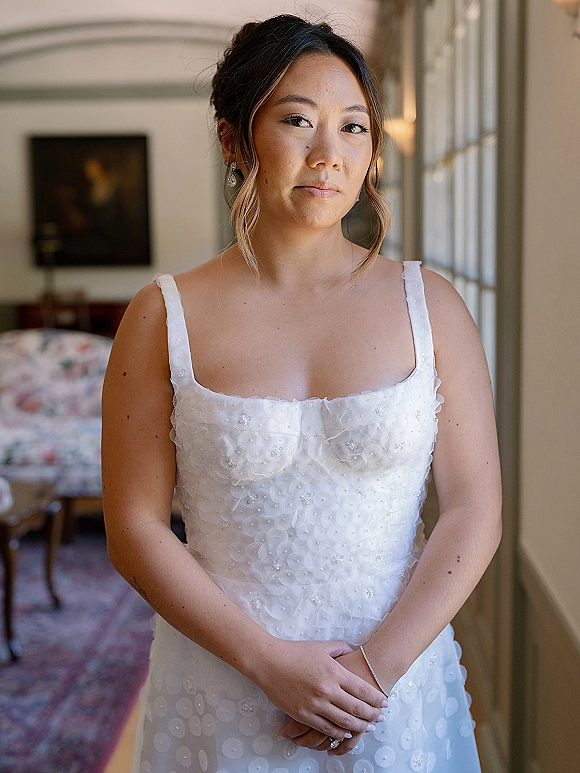 Bridal portrait of a bride getting ready in a spaghetti-strap beaded floral dress, hands clasped by window light in a bedroom interior