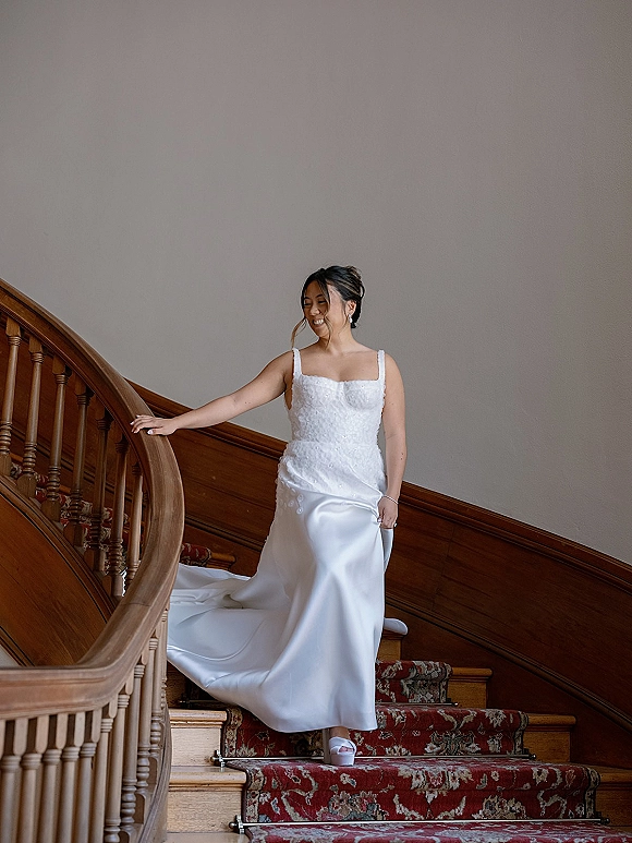 Bridal portrait of a bride on staircase, holding her satin skirt to descend in a lace bodice gown with a long train on a wood stairway