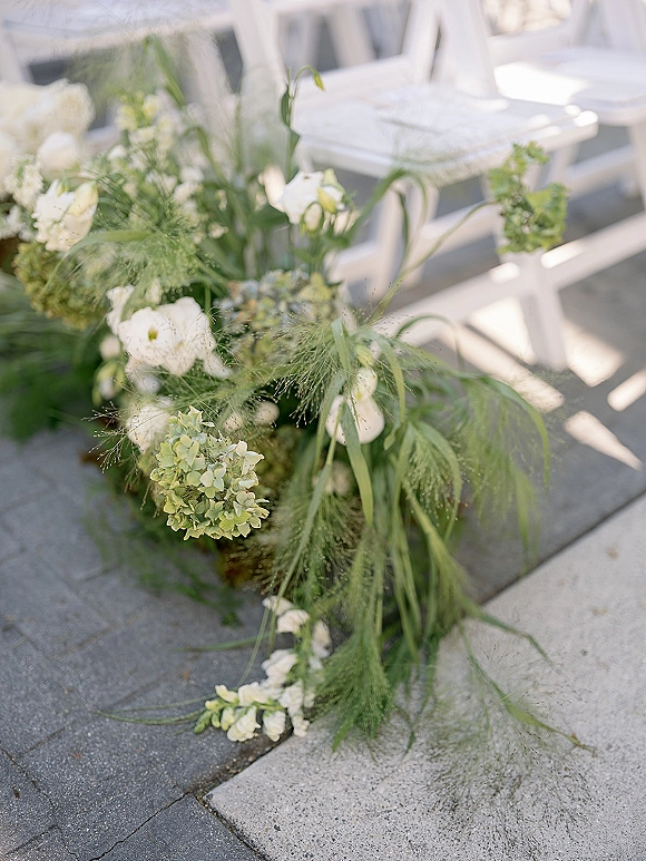 Ceremony aisle flowers in a low white and green floral arrangement with hydrangea and roses beside white folding chairs on stone pavement