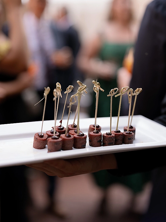 Wedding appetizers on a white platter, passed hors d'oeuvres on cocktail skewers held by a server amid a cocktail hour crowd