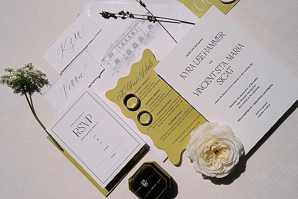 Invitation suite flat lay with black and white wedding invitations, RSVP and place cards, rings, ring box, and white flower on linen
