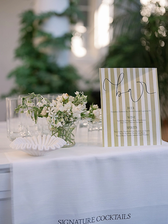 Wedding bar signage with a signature cocktails sign in calligraphy on a striped menu, set on a white tablecloth with florals outdoors