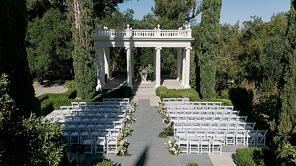 Ceremony setup for an outdoor wedding ceremony with white folding chairs and greenery-lined aisle florals leading to a pergola altar under blue sky