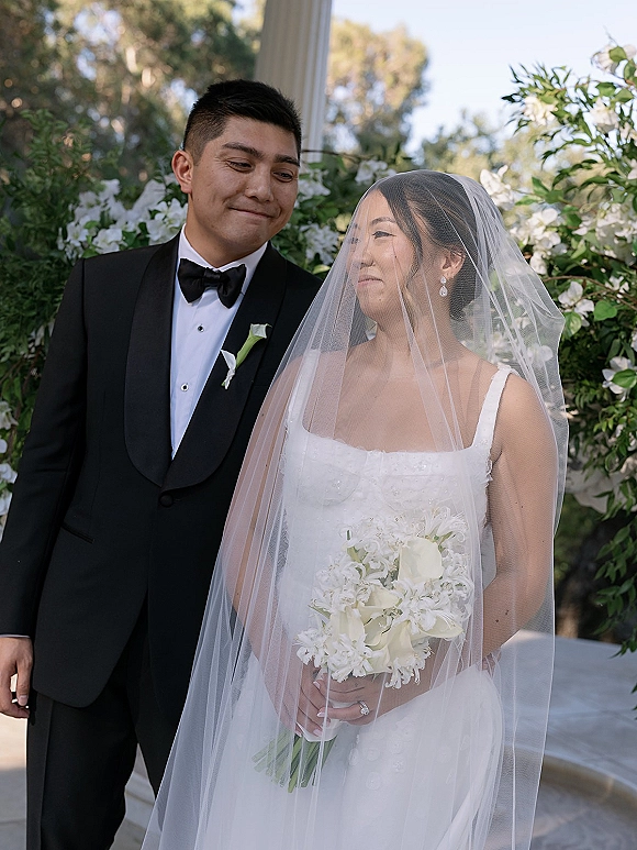 Couple portrait of bride and groom portrait with veil and white calla lily bouquet, posing in a garden by greenery and white flowers
