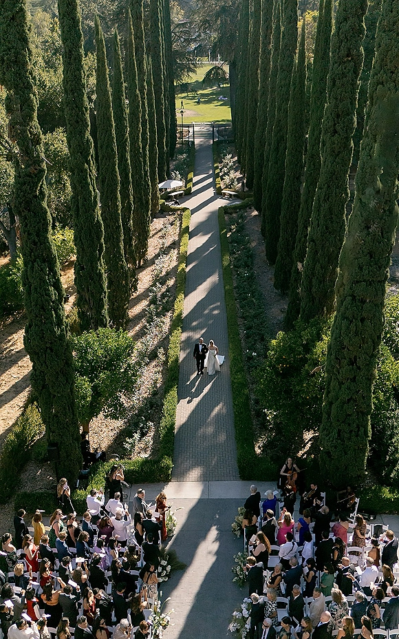 Wedding processional with bride walking down aisle between white ceremony chairs and floral arrangements on a sunlit tree-lined garden path
