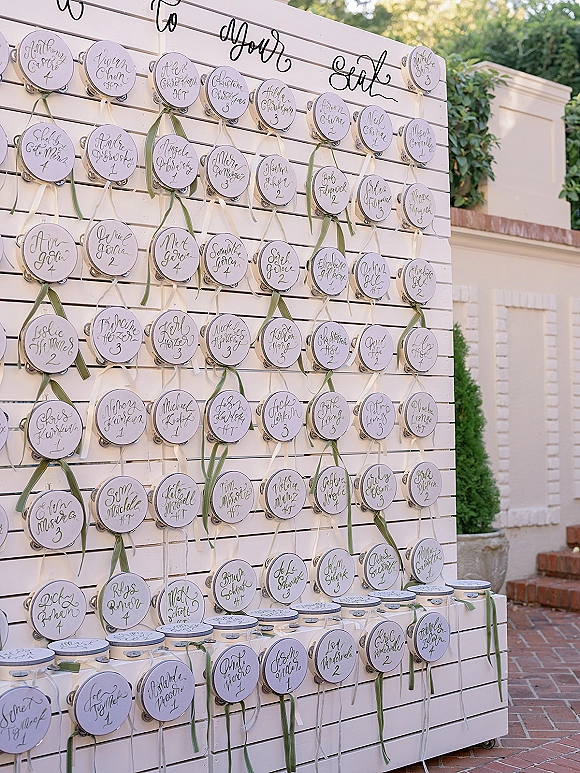 Wedding escort display with wedding escort cards on a white slat wall, featuring calligraphy round tags, ribbons, and metal tins outdoors