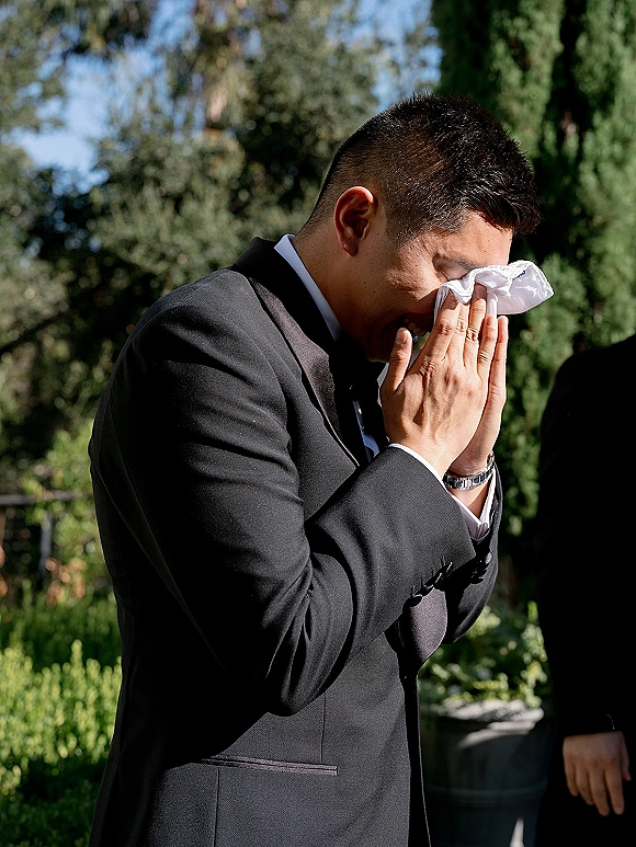 Groom portrait wiping tears with a handkerchief in a black tuxedo and bow tie, sunlight filtering through garden trees behind him
