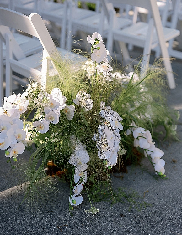 Ceremony aisle florals with white orchid aisle arrangement, baby’s breath and greenery clusters lining white folding chairs on outdoor pavement