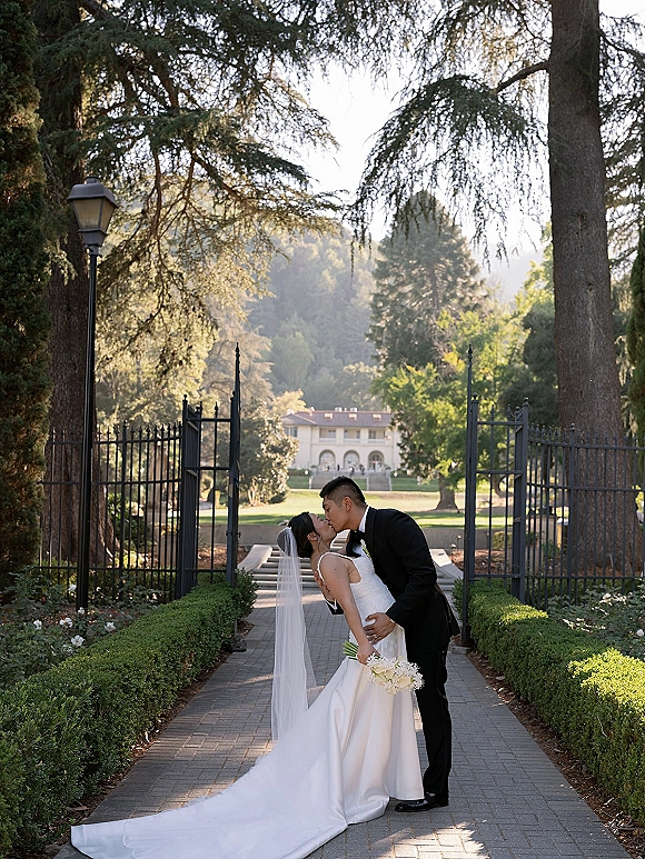 Wedding kiss in a romantic dip as the groom holds the bride in a strapless satin dress and veil on a garden walkway by iron gates