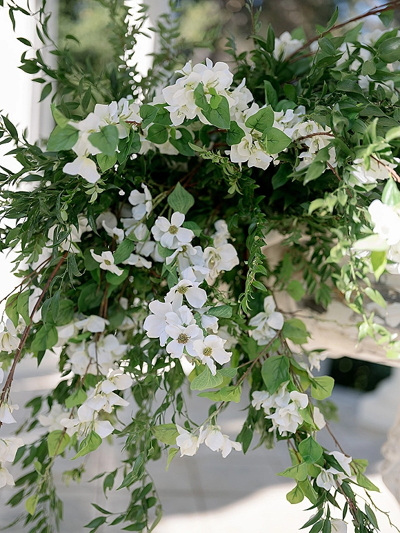 Wedding floral arrangement of white and green wedding flowers with leafy vines and branches set against a sunlit white wall outdoors