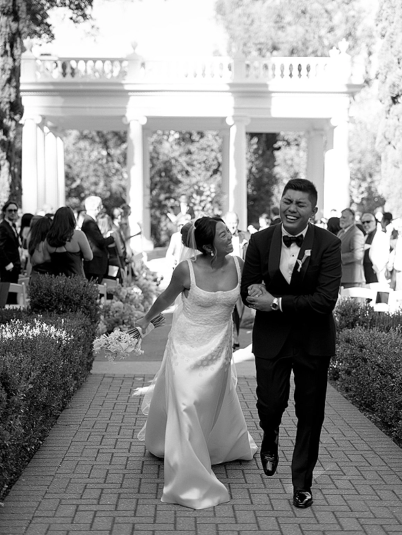 Wedding recessional with bride and groom walking, smiling with bouquet and veil as guests line a brick garden aisle by white colonnade pavilion