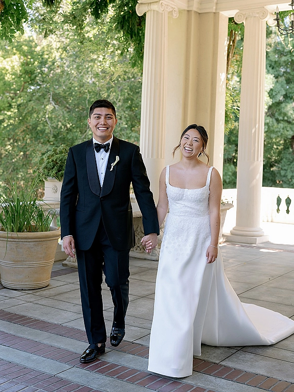 Couple portrait of bride and groom walking hand in hand on a columned terrace, her satin square neckline dress and his black tuxedo by greenery