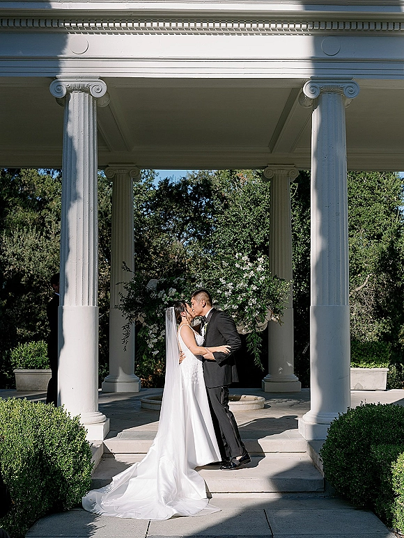 Wedding kiss portrait of bride and groom kiss under a white column pavilion, her long veil and train draped down stone steps