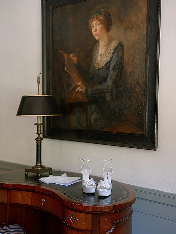Wedding shoes with bridal shoes flatlay styling, white platform ankle-strap heels on a leather desk blotter beside stationery on an antique writing desk