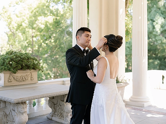 First look moment as groom seeing bride in a strapless gown, adjusting her updo on a stone terrace with white columns and greenery
