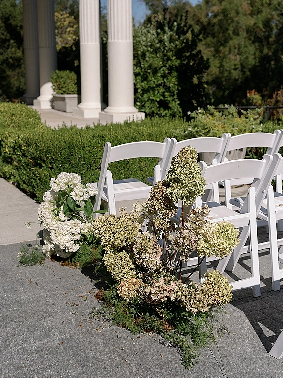 Ceremony aisle decor with hydrangea aisle flowers beside white folding chairs, greenery garland along a stone garden walkway with columns