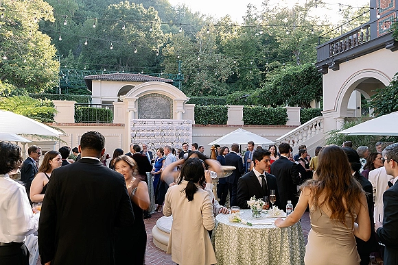 Cocktail hour with wedding cocktail hour guests mingling at high-top tables under string lights in an ivy-lined brick courtyard with arches