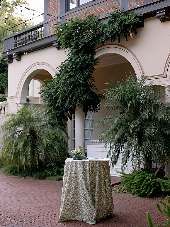Wedding guestbook table with a guestbook and pen on a patterned tablecloth, small floral arrangement and string lights in an ivy courtyard.