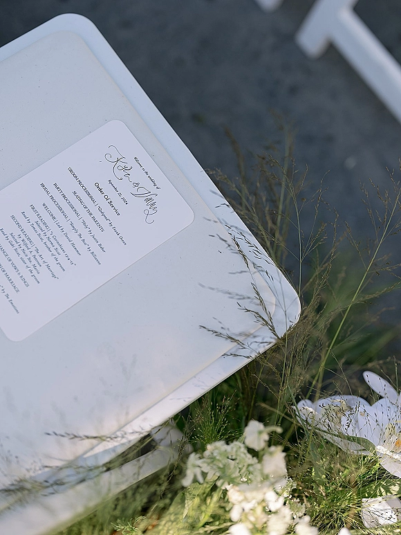 Wedding ceremony program resting on a white chair with greenery accents and wild grass, set against asphalt pavement outdoors