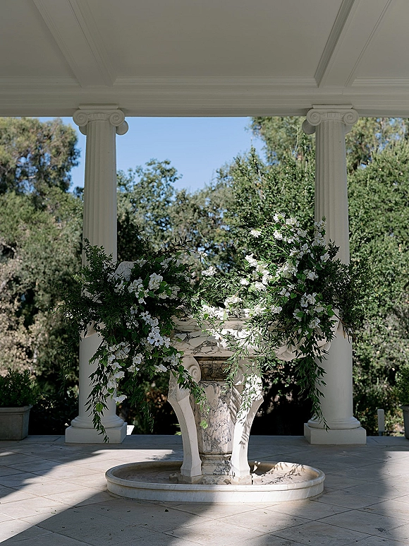Wedding floral installation with white flowers and greenery garland draped over a marble fountain and columns under a pavilion ceiling