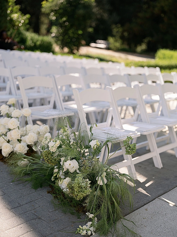 Ceremony seating with outdoor ceremony chairs in neat white folding chair rows, framed by low white rose and hydrangea aisle flowers in a sunlit garden