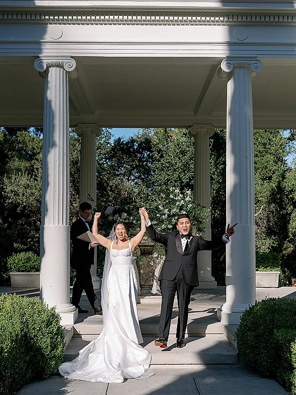 Recessional moment as bride and groom exit just married, hands raised on stone steps beneath a floral arch at a columned pavilion