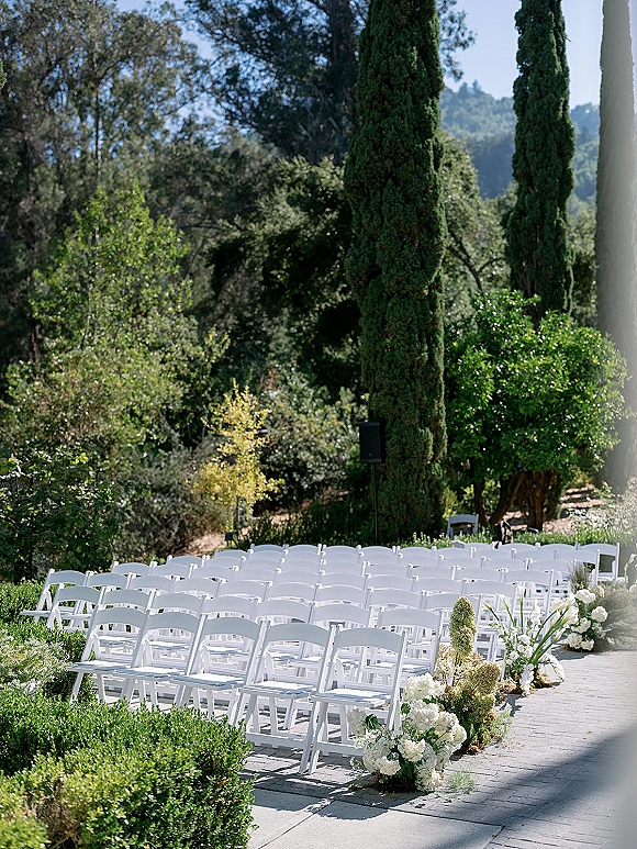 Outdoor ceremony setup with white folding chairs lining a stone patio aisle, hydrangea and greenery clusters, cypress trees beyond