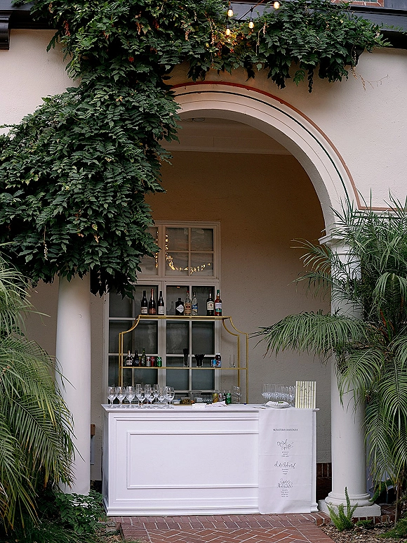 Wedding bar setup with a white counter and liquor bottles, stemware, and menu sign in an arched alcove on a brick patio