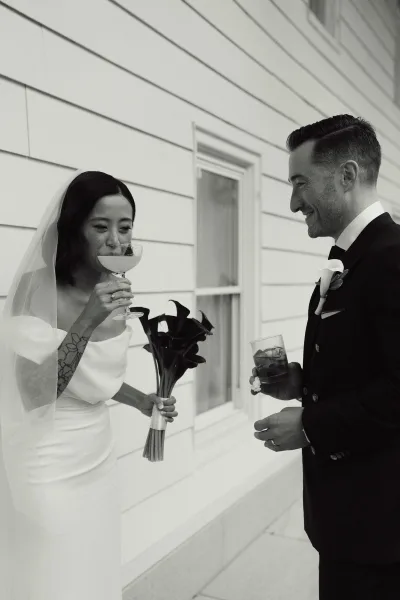 Couple portrait in a black and white wedding portrait style, bride sipping champagne beside groom holding whiskey by a white clapboard wall.