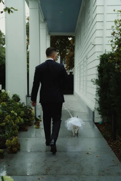 Groom and dog strolling on a leash, pup in a wedding dress with veil accent, along a sidewalk by a white house porch and shrubs