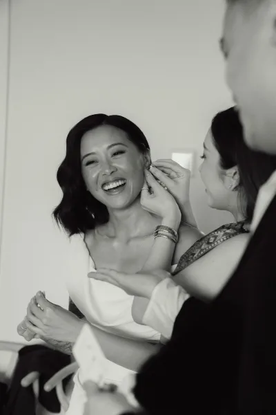 Bride getting ready as a bridesmaid helps her put on drop earrings, smiling in a strapless dress with bracelets in a simple room